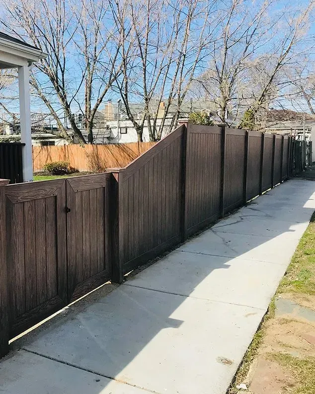 Brown privacy fence bordering a concrete sidewalk. Trees and houses in the background under a blue sky.