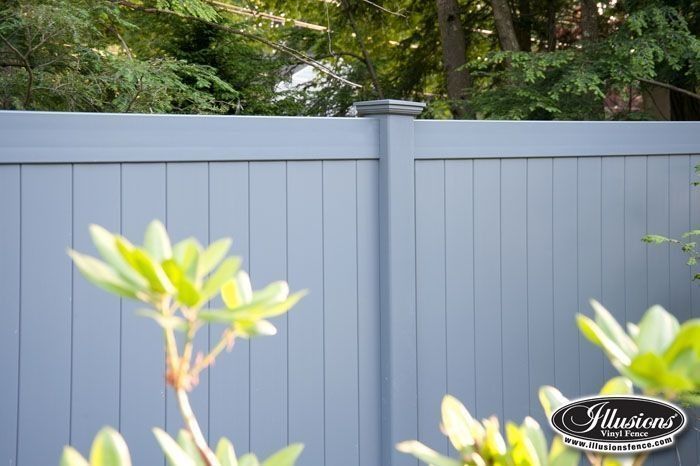 Gray vinyl fence with vertical planks and a dark gray post cap, with blurred greenery in the foreground and background.
